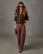Woman in brown knit button cardigan, wide-leg chocolate trousers, sunglasses, gold cuff and pointed heels holding a clutch