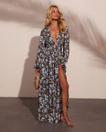 Woman wearing olive floral maxi dress with blue print, deep V-neck, side slit, long sleeves, nude heels, holding woven clutch