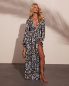 Woman wearing olive floral maxi dress with blue print, deep V-neck, side slit, long sleeves, nude heels, holding woven clutch