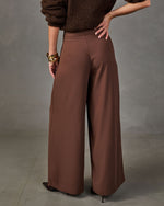 Back view of high-waisted brown wide-leg trousers styled with a chunky knit sweater, gold bangles and pointed heels