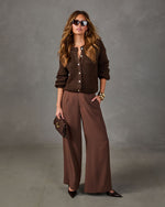 Woman in brown button-up knit cardigan, wide-leg brown trousers, sunglasses, gold bangles and pointed heels holding a clutch