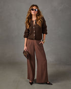 Woman in brown button-up knit cardigan, wide-leg brown trousers, sunglasses, gold bangles and pointed heels holding a clutch