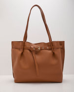 A brown pebbled leather tote bag with dual shoulder straps, a front strap detail, and gold-tone hardware, photographed against a neutral light gray background.