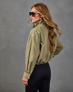 Woman wearing olive green zip-up cropped jacket with black leggings, gold hoop earrings, and aviator sunglasses, back view