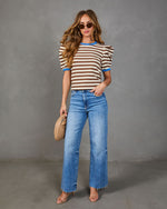 Woman in Marcine Striped Puff Sleeve Top, light-wash jeans, and beige sandals holds a tan clutch, showcasing a relaxed yet stylish ensemble.