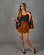 A woman models a stylish outfit consisting of a black ribbed tank top, a brown velvet mini skirt, a matching brown blazer draped over her shoulders, and a black woven handbag, set against a gray studio backdrop.