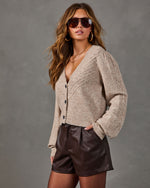 Woman in a beige pointelle button-front cardigan with puff sleeves, brown faux leather shorts, aviator sunglasses and gold hoops