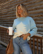 A woman stands on a wooden deck in front of a log cabin wall, wearing a light blue crewneck sweater with a white tree design, blue jeans, and holding a white coffee cup.