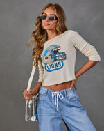 A woman modeling a beige long-sleeve crop top with a blue and silver football helmet graphic and the word 'LIONS' on the front, paired with light blue jeans and sunglasses against a gray studio background.