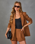 Woman wearing camel brown blazer and shorts set with black bodysuit, cat-eye sunglasses, and woven leather clutch bag