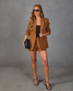 Woman wearing camel brown blazer shorts suit set with black bodysuit, strappy heels, woven clutch, and cat-eye sunglasses