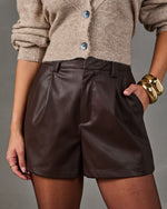 Brown faux leather pleated high-waisted shorts styled with a beige knit cardigan and chunky gold cuff bracelets