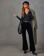 A woman models a stylish outfit consisting of a black ribbed long-sleeve henley top, black wide-leg velvet pants, a black beret, black sunglasses, a black quilted shoulder bag, and a textured gray overcoat draped over her arm, set against a neutral gray studio backdrop.