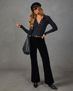 A woman models a black ribbed long-sleeve polo shirt with button details, paired with black flared pants, a black beret, sunglasses, a black quilted shoulder bag, and black boots, set against a textured gray studio background.