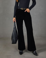A model wearing black wide-leg corduroy pants, a black ribbed long-sleeve top, and black pointed-toe ankle boots, holding a black quilted shoulder bag against a gray studio backdrop.