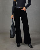 A model wearing black wide-leg corduroy pants, a black ribbed long-sleeve top, and black pointed-toe ankle boots, holding a black quilted shoulder bag against a gray studio backdrop.