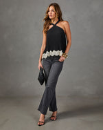 A woman models a black halter-neck top with a white lace hem, paired with dark gray flare jeans, black strappy heels, and a small black clutch, set against a textured gray studio backdrop.