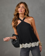 A woman modeling a black halter-neck pleated top with a cream lace hem, paired with dark gray jeans and holding a black quilted handbag against a textured gray studio background.