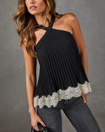 A woman wearing a black halter-neck pleated top with a cream lace hem, paired with dark gray jeans, against a neutral gray studio background.