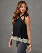 A woman modeling a black halter-neck pleated top with a white lace hem, paired with gray jeans, against a neutral gray studio background.