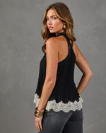 A woman modeling a black sleeveless halter top with a white lace hem, paired with gray jeans, against a textured gray studio background.