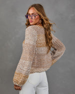 Woman in a beige and tan chunky knit sweater with white jeans and tortoiseshell glasses, looking back over her shoulder