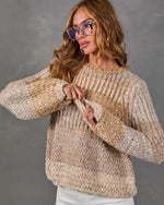Woman wearing a chunky beige and caramel marled knit sweater with tortoiseshell glasses and white pants against a gray backdrop