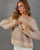 Woman wearing a chunky beige and caramel marled knit sweater with tortoiseshell glasses and white pants against a gray backdrop