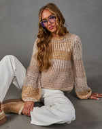 Woman seated in a beige striped chunky knit sweater, white wide-leg pants, tortoiseshell glasses and tan platform slippers