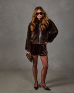 Woman in brown faux fur bomber jacket, velvet shorts, sheer tights and slingback heels holding a mini clutch, aviator sunglasses