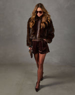 Woman in brown faux fur bomber jacket, velvet pleated shorts, sheer tights, aviator sunglasses and pointed pumps with mini bag