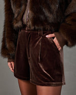 Brown velvet pleated high-waisted shorts styled with a faux fur bomber jacket and sheer tights, hand in pocket
