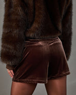 Model in brown velvet shorts with back welt pockets, sheer tights and a cropped dark brown faux fur jacket, back view