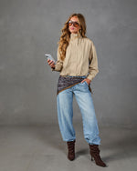 Woman in tan zip jacket, paisley wrap, light wash wide-leg jeans, dark brown slouch boots, oversized sunglasses, holding phone