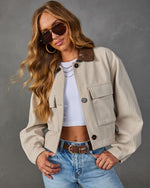 A woman models a cropped beige utility jacket with a brown collar, worn over a white crop top and blue jeans, against a textured gray studio background.