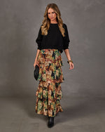 A woman models a long black knit sweater paired with a tiered, floral-printed maxi skirt in earthy tones of green, brown, and beige. She is standing against a textured gray studio backdrop, holding a small black quilted clutch.