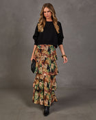 A woman models a long black knit sweater paired with a tiered, floral-printed maxi skirt in earthy tones of green, brown, and beige. She is standing against a textured gray studio backdrop, holding a small black quilted clutch.