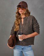 Woman wearing a brown boucle button-up cardigan, suede cap, tinted sunglasses and light-wash jeans, holding a coffee and woven clutch