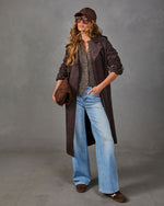 Woman in a brown suede trench coat, gray knit cardigan vest, wide-leg light wash jeans, suede cap, and woven clutch
