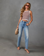 Woman wearing brown striped crop tank top, light wash straight-leg jeans, nude heeled sandals, and tinted sunglasses holding woven bag