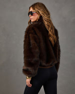Woman in a cropped brown faux fur jacket with black leggings, aviator sunglasses and gold jewelry, seen from behind