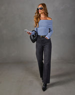 Woman in blue striped off-the-shoulder sweater, black wide-leg jeans, woven clutch, sunglasses and black ankle boots
