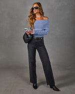 Woman in blue striped off-the-shoulder sweater, black wide-leg jeans, gold buckle belt, woven bag and pointed ankle boots