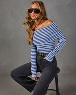 Woman in blue and black striped off-the-shoulder sweater, gray wide-leg jeans and black sunglasses, seated on a stool