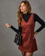Woman in a rust brown suede mini pinafore dress over a black mock neck top, with a black woven handbag and gold bangles