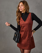 Woman in a rust brown suede mini pinafore dress over a black mock neck top, with a black woven handbag and gold bangles