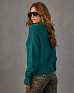 Woman in a teal green knit sweater with ribbed hem and cuffs, styled with leopard print jeans and black aviator sunglasses