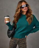 Woman in a teal mock neck knit sweater, leopard print jeans, black woven bag and sunglasses holding a takeaway coffee cup
