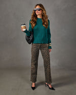 Woman in emerald green knit sweater, leopard print jeans and black heels holding a coffee cup and woven black bag