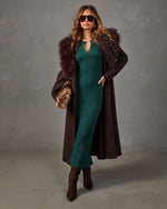 Woman in a green ribbed knit midi dress, long brown coat with faux fur collar, suede ankle boots and aviator sunglasses
